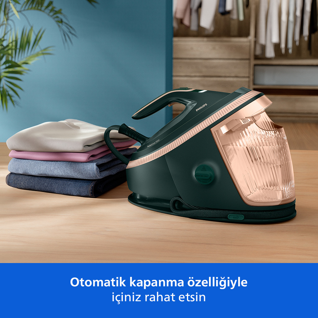 Koyu yeşil buharli ütü, ahşap masa üzerinde yığılı giysilerin yanında.