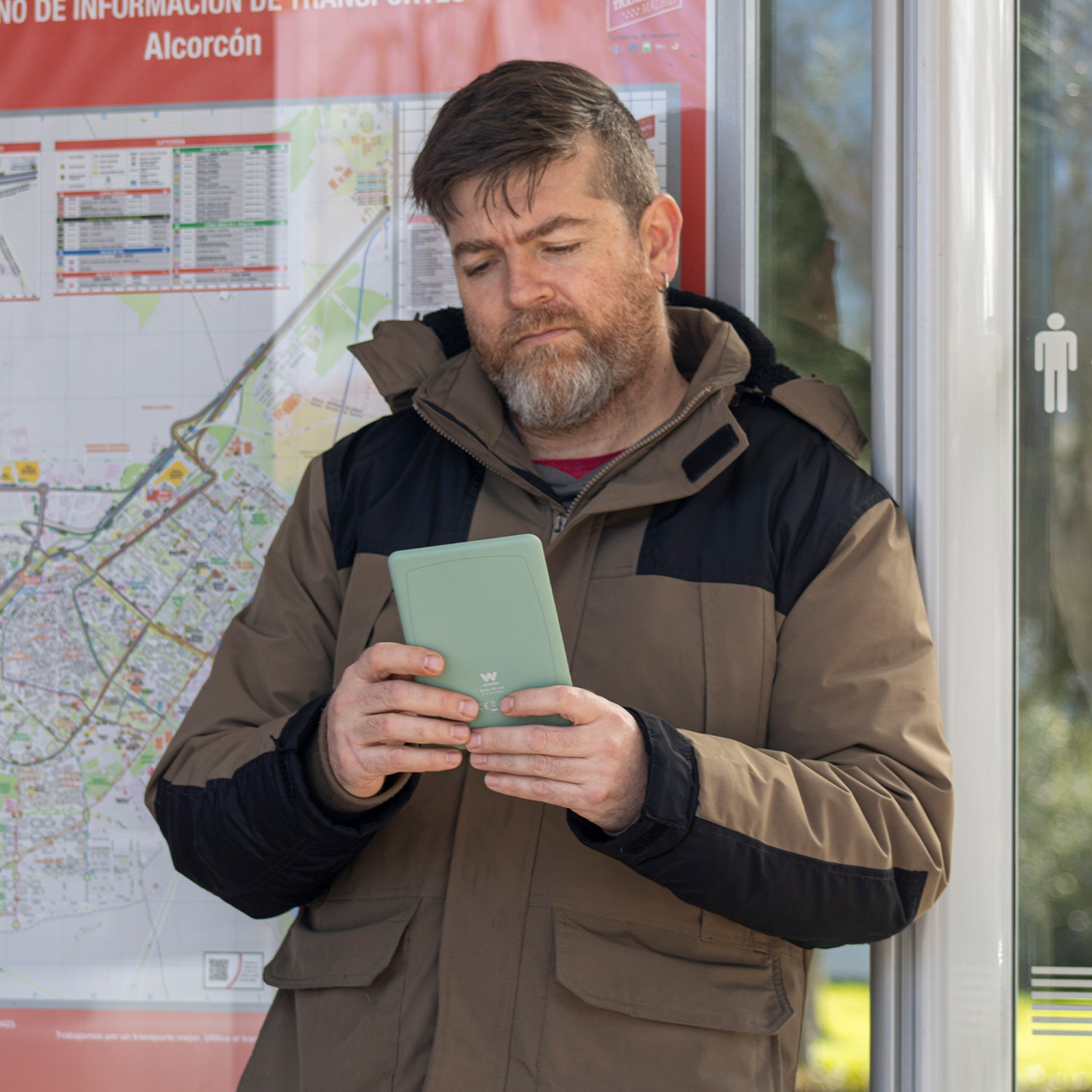 Un hombre con barba sostiene un dispositivo verde claro con un abrigo marrón junto a un mapa.