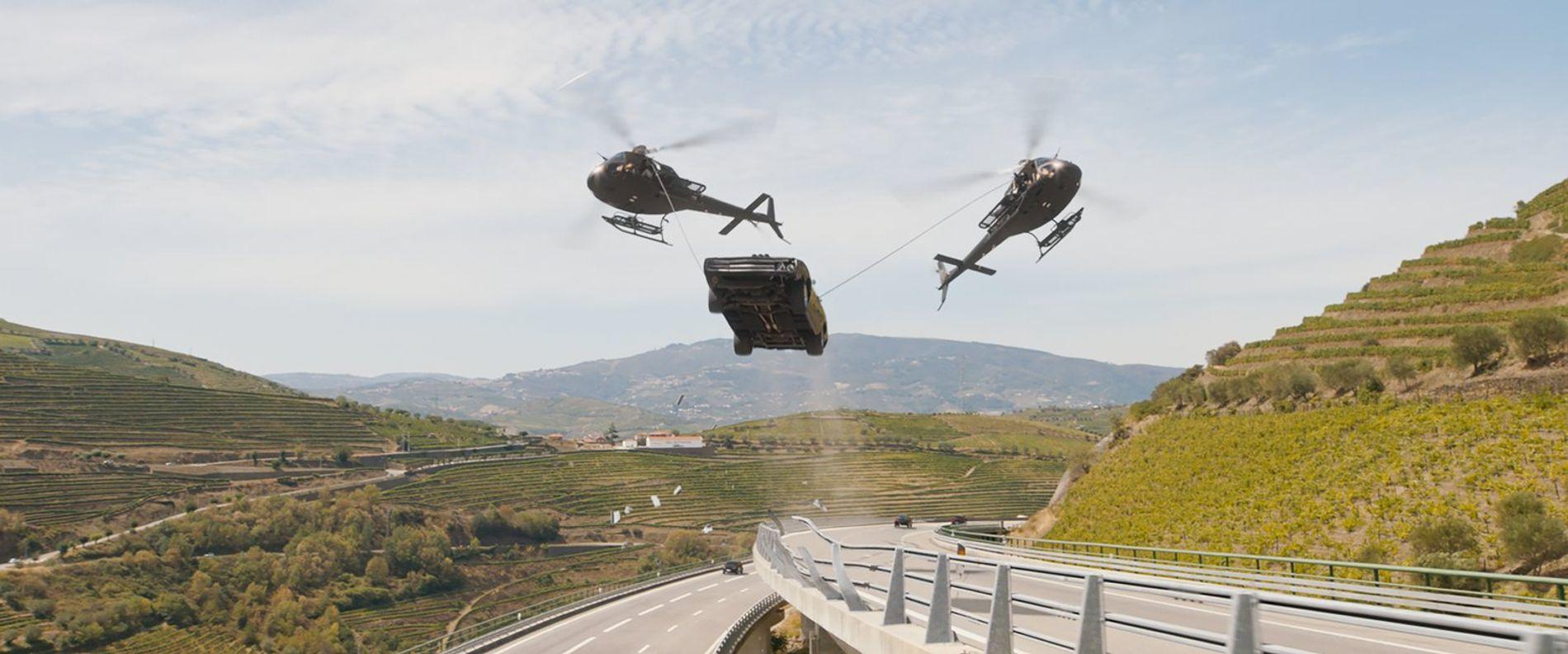 Zwei Hubschrauber hängen ein Auto über einer Autobahn in einer bergigen Landschaft.