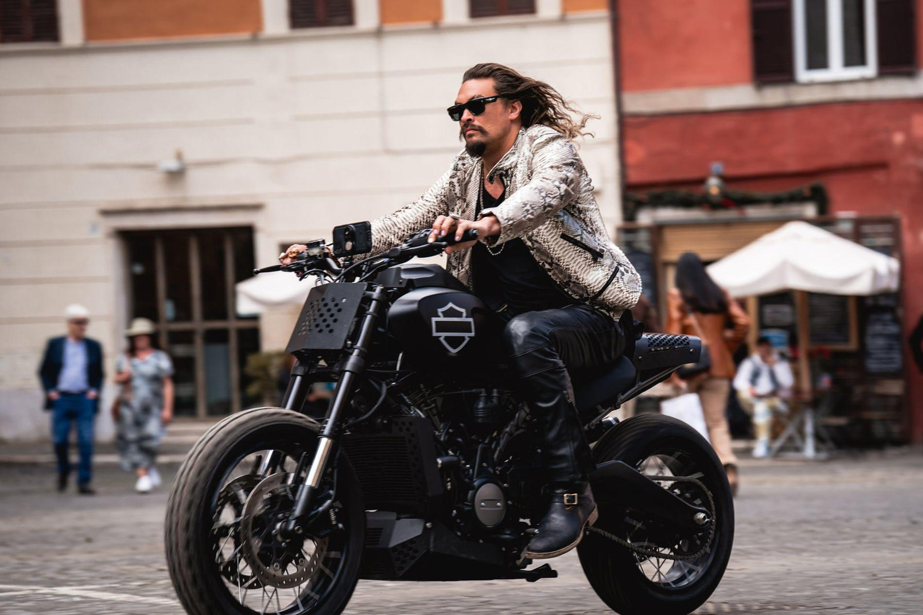 Mann mit langen Haaren fährt ein schwarzes Motorrad in einer Stadtstraße.