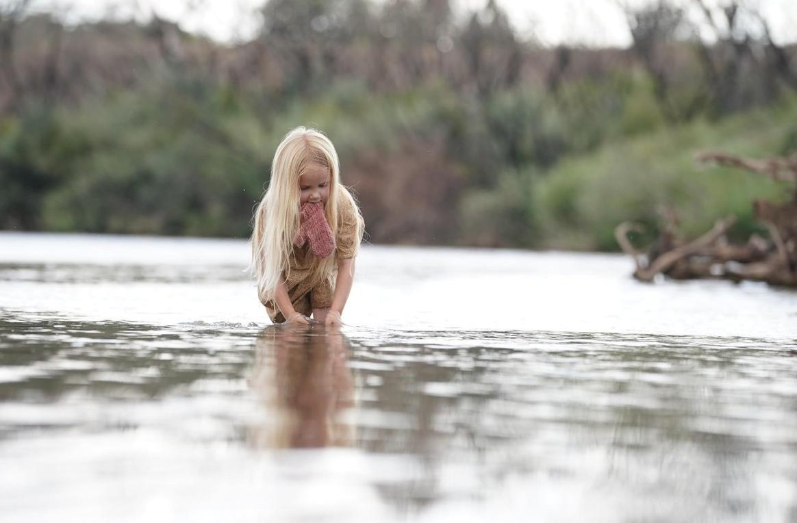 Ein kleines Mädchen mit langen blonden Haaren watet im Wasser und hält ein rosafarbenes Kleidungsstück.