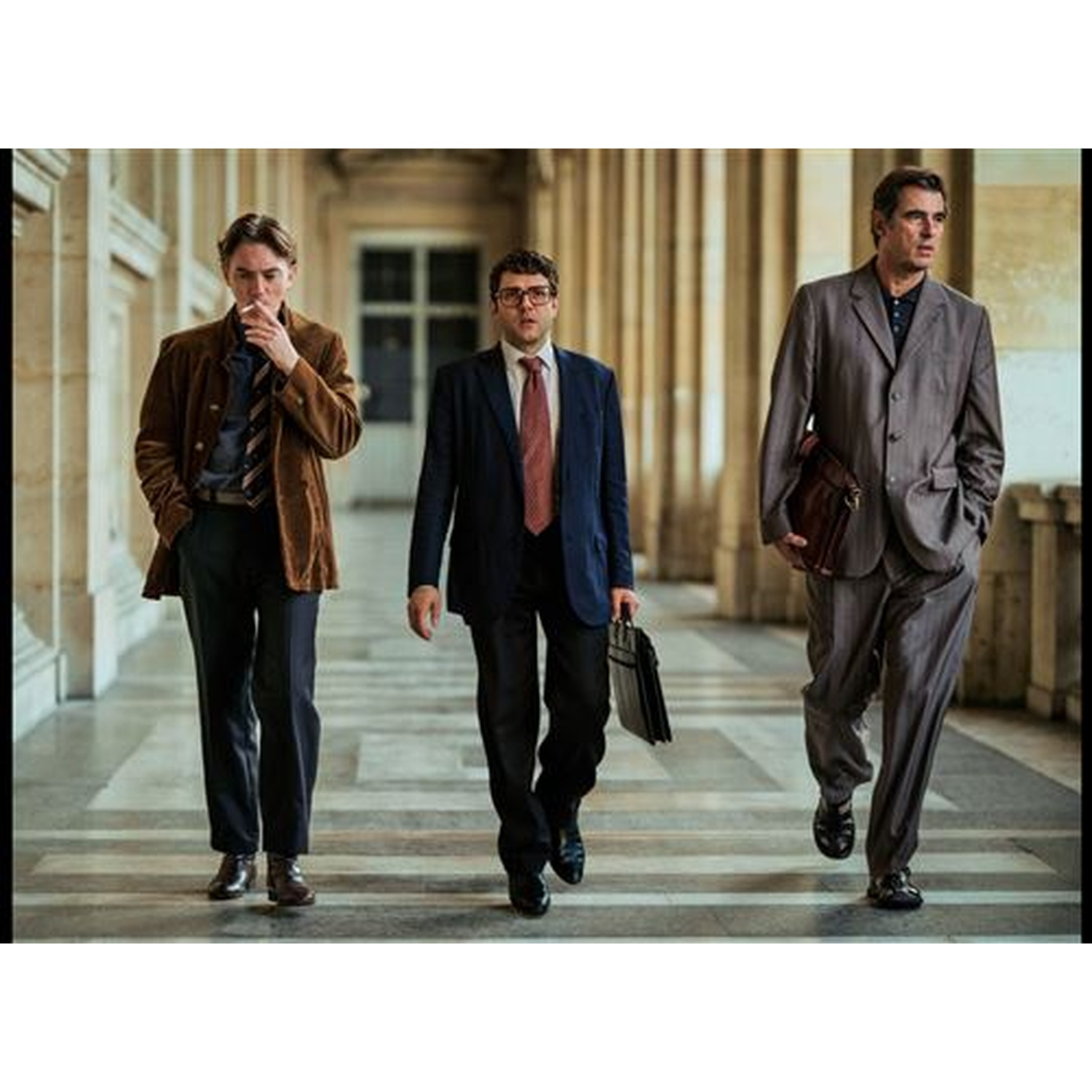 Drie mannen in pak lopen door een gang. Eén rookt, de anderen dragen aktetassen.