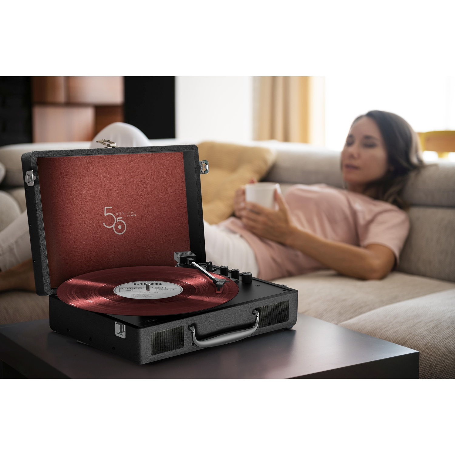 Een platenspeler met rode vinylplaat staat op tafel, vrouw ontspant op de bank op achtergrond.