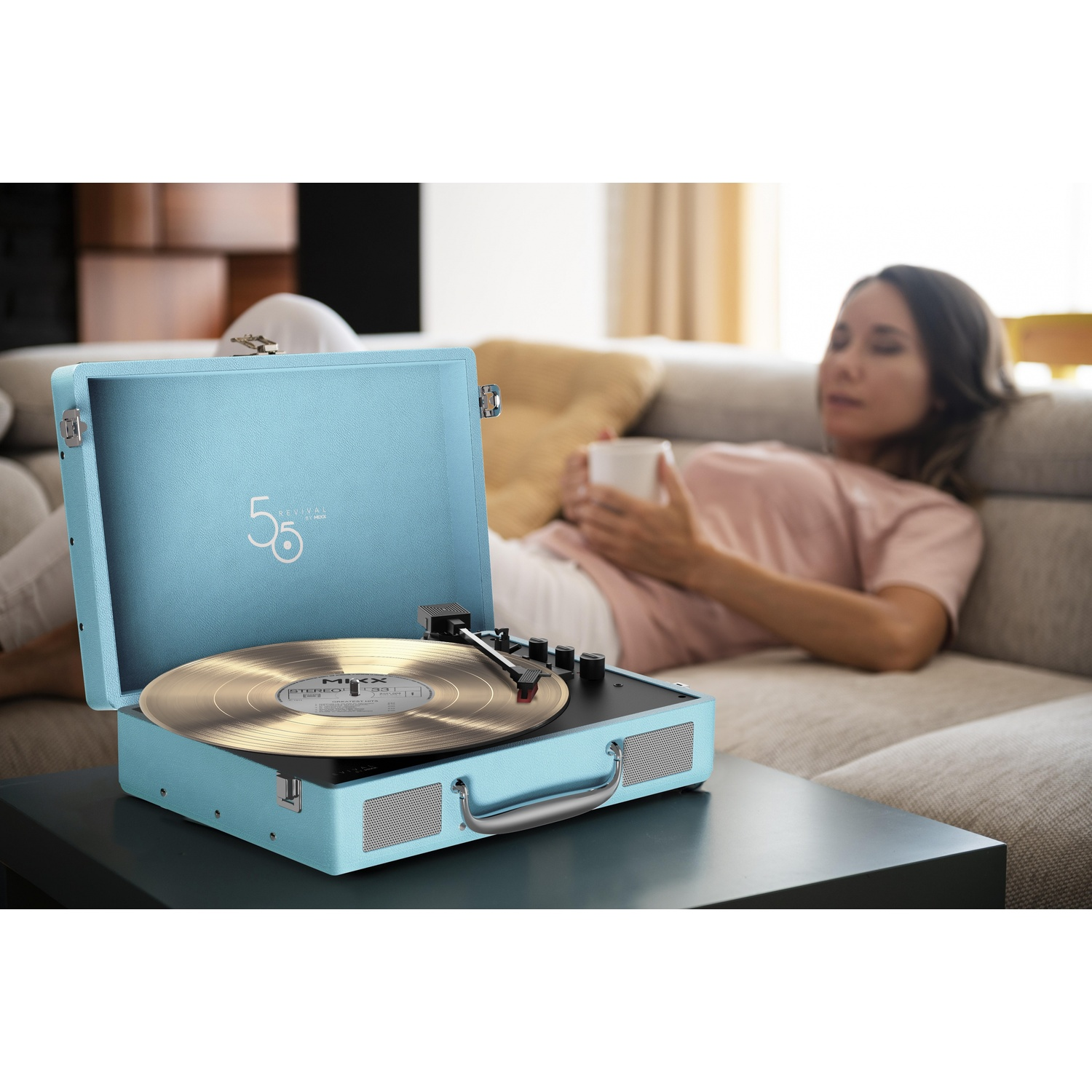 Een lichtblauwe platenspeler met vinylplaat op tafel, een vrouw drinkt.