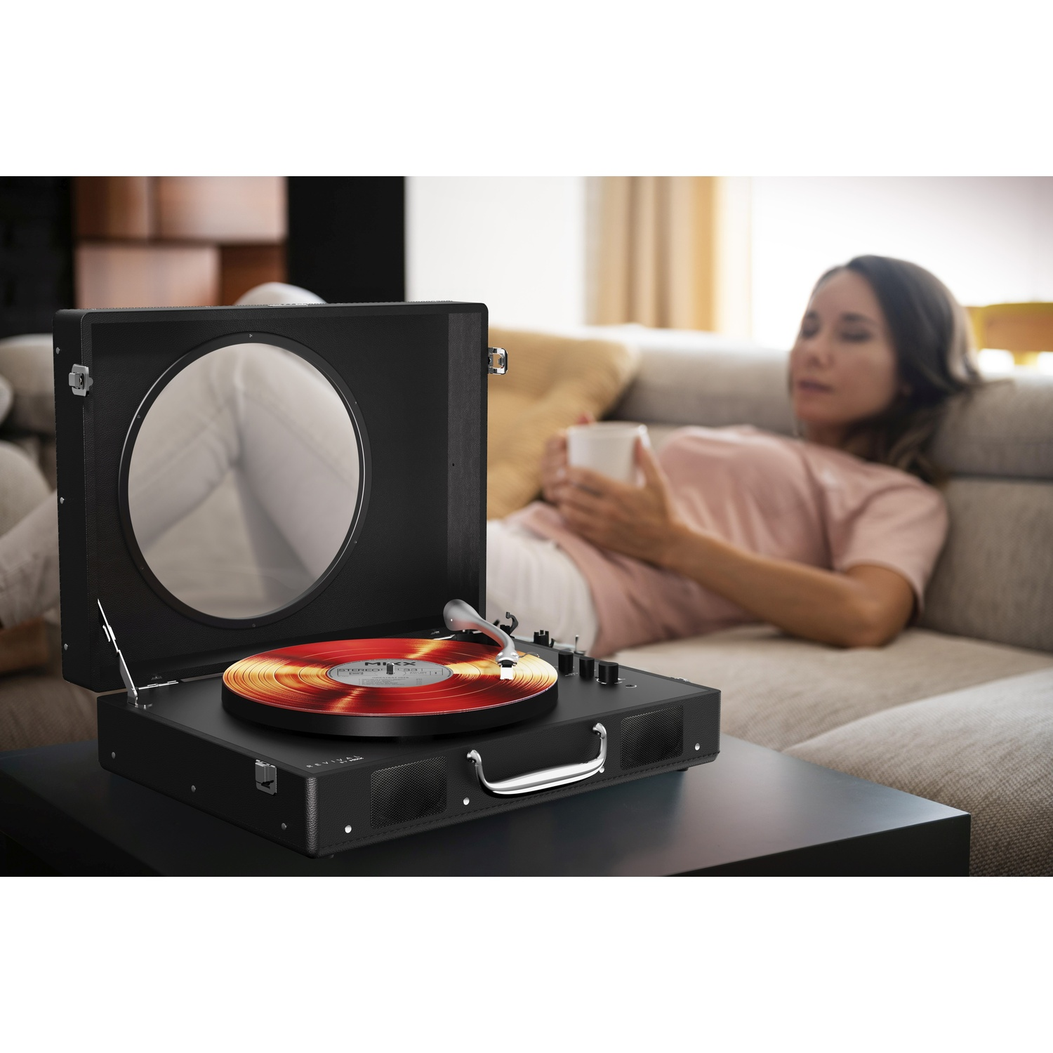 Une platine vinyle joue un disque rouge et doré, une femme se détend sur un canapé.