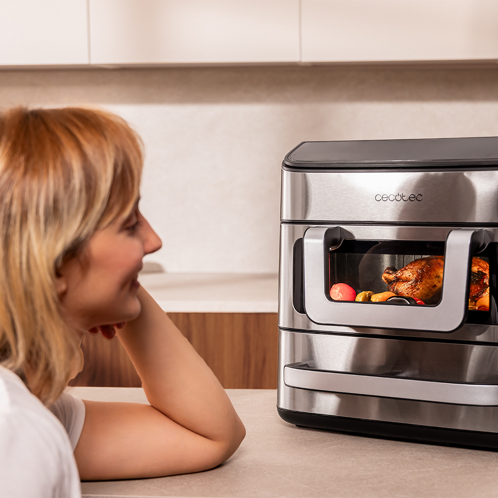 Una mujer mira un pollo en un horno. Se ven guarniciones como zanahorias.
