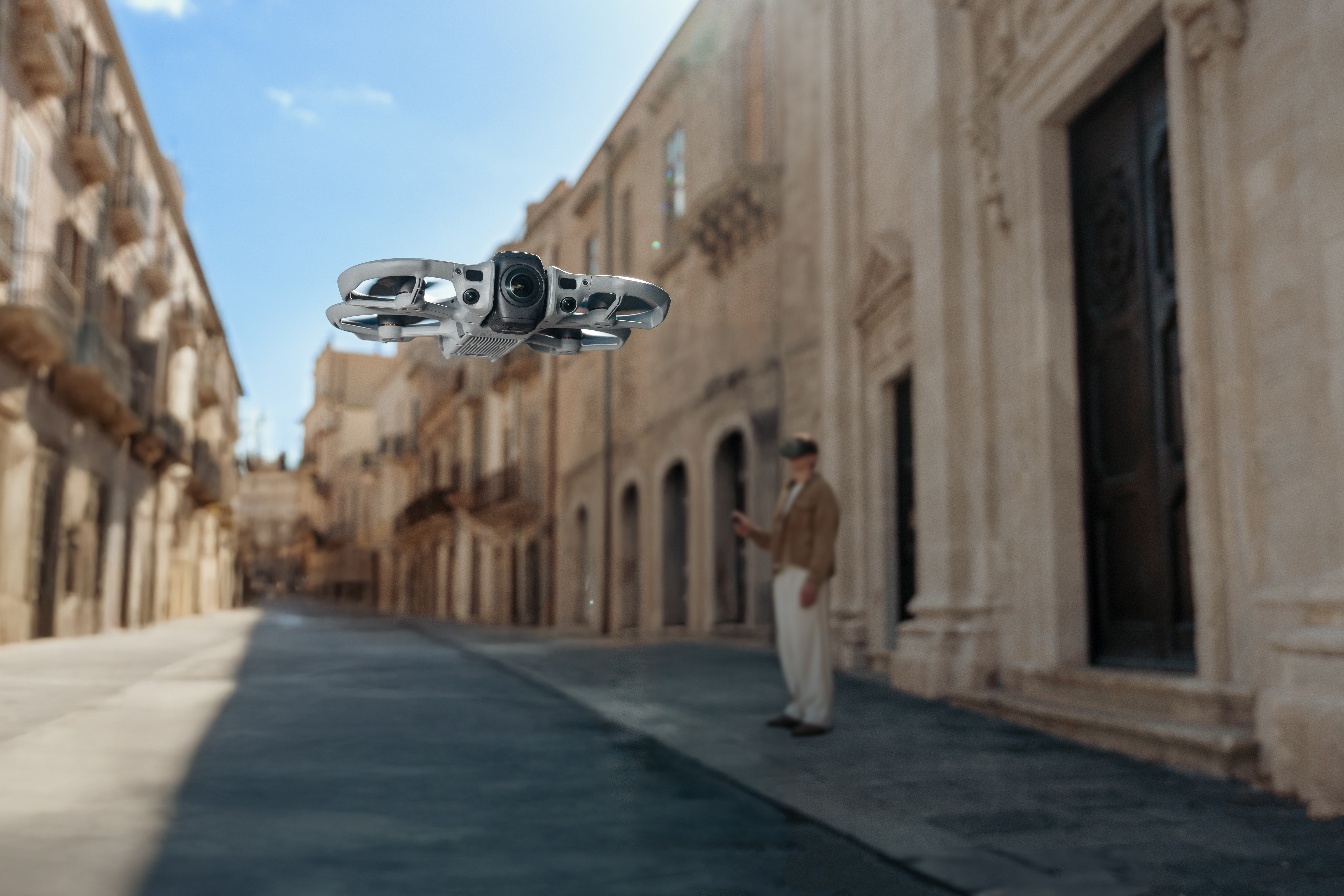 Eine weiße Drohne fliegt über eine Straße mit Gebäuden. Ein Mann mit VR-Brille steht im Hintergrund.
