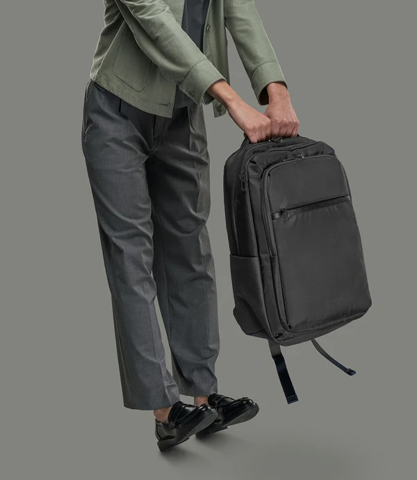 Persona con pantalón gris y chaqueta verde sostiene una mochila negra.