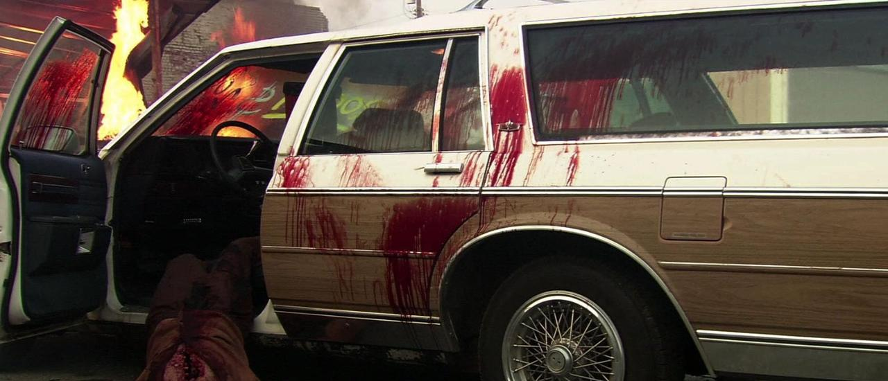 Ein offenes Auto mit viel Blut, Flammen im Hintergrund.