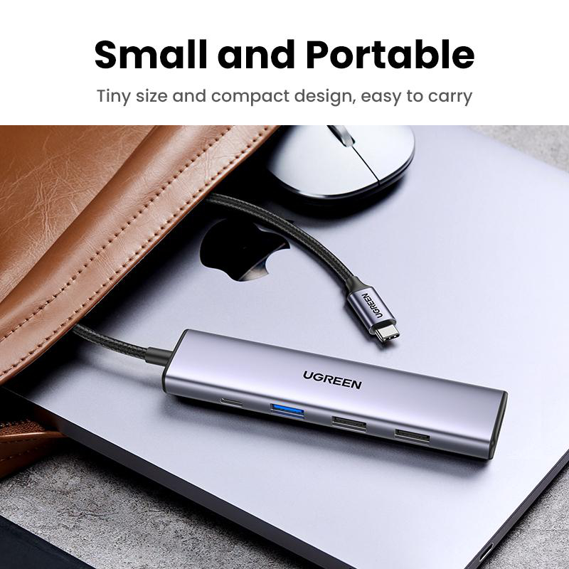 Ein silberner USB-C-Hub liegt auf einem Laptop, daneben eine Maus und eine braune Tasche.