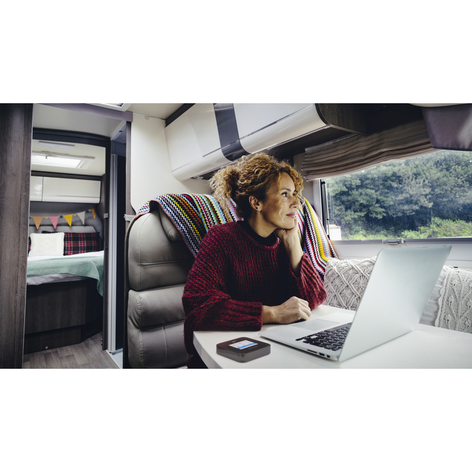 Vrouw in camper met laptop en klein apparaat op tafel.