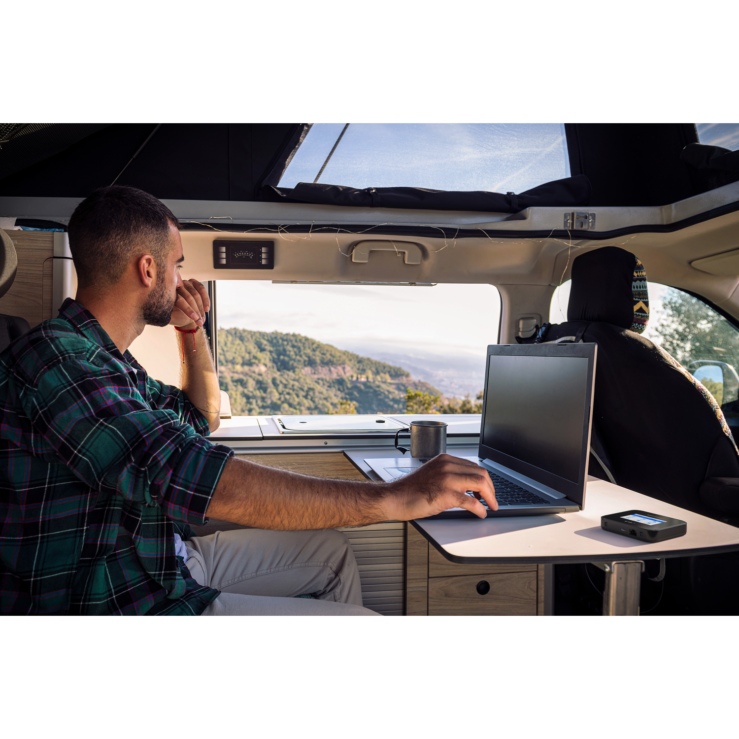 Man in camper werkt aan laptop, met uitzicht op bergen en lucht.