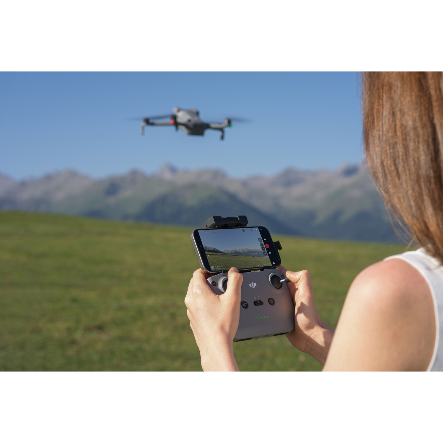 A woman controls a drone with a remote in a grassy field, mountains in the distance.
