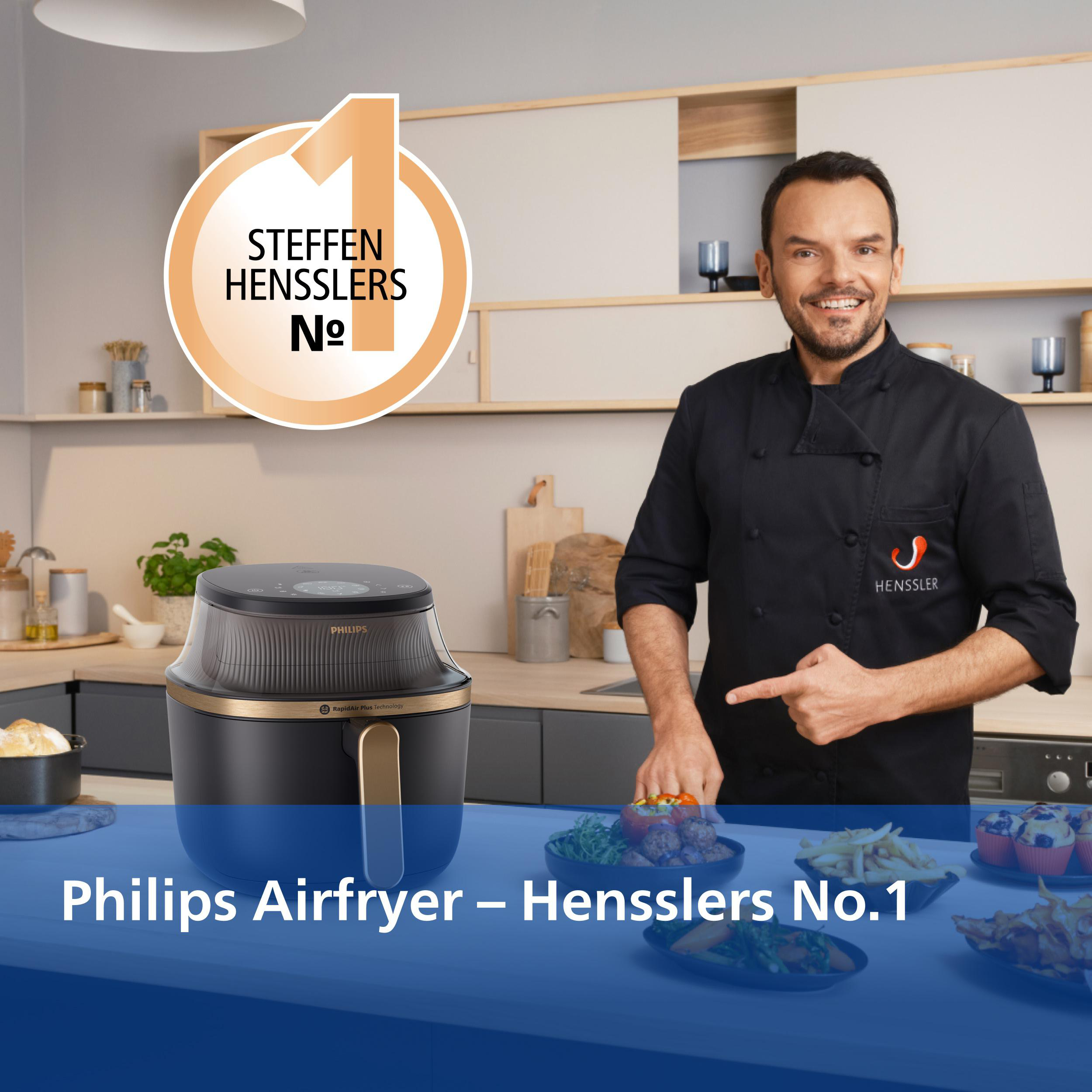 Ein Mann im schwarzen Kochkittel zeigt auf einen Philips Airfryer neben Essen.