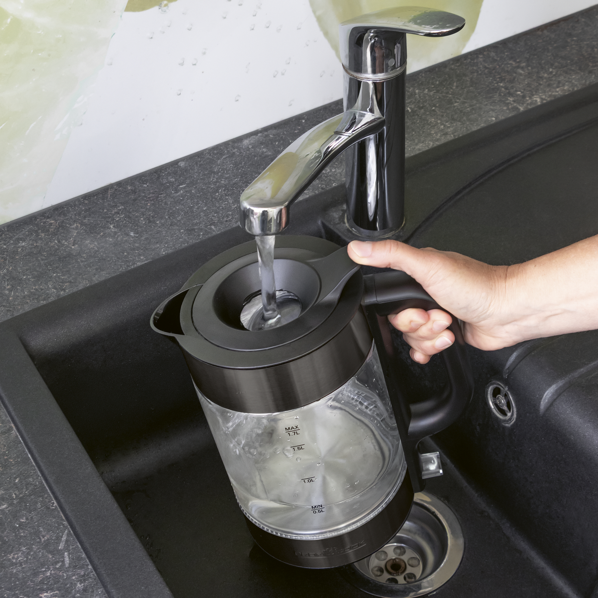Eine Hand füllt einen transparenten Wasserkocher mit Wasser unter einem Wasserhahn.
