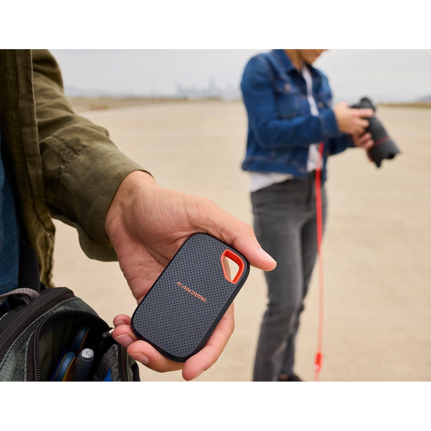Una mano tiene un disco SanDisk nero con anello arancione. Sullo sfondo, una persona con macchina fotografica.