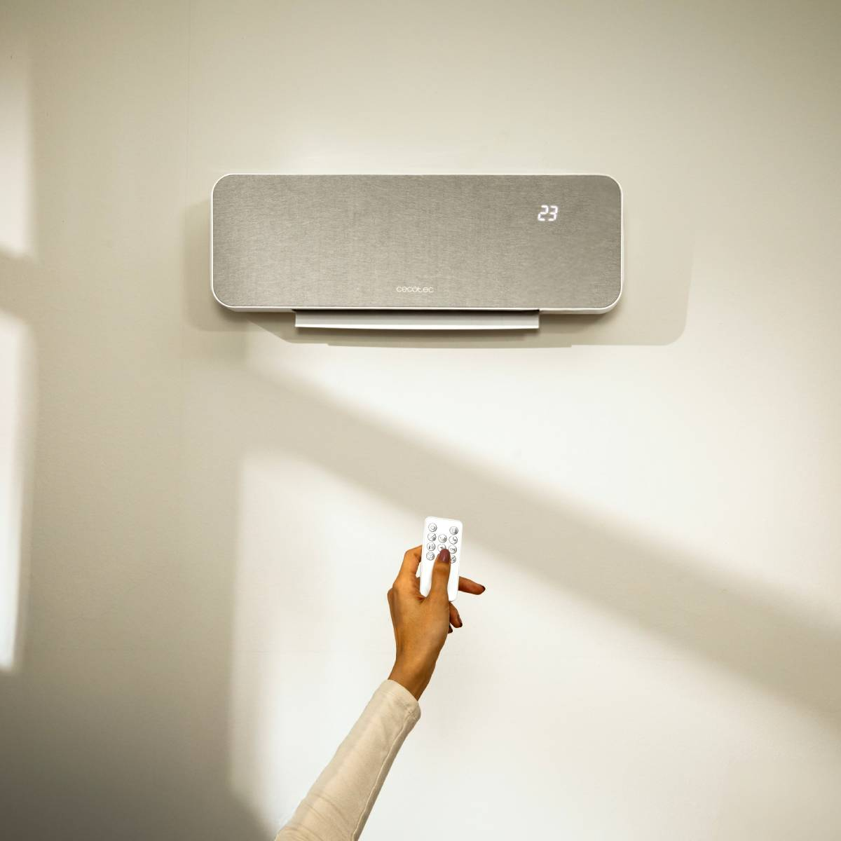 Una mano sostiene un mando a distancia frente a un aire acondicionado de pared que muestra 23°C.