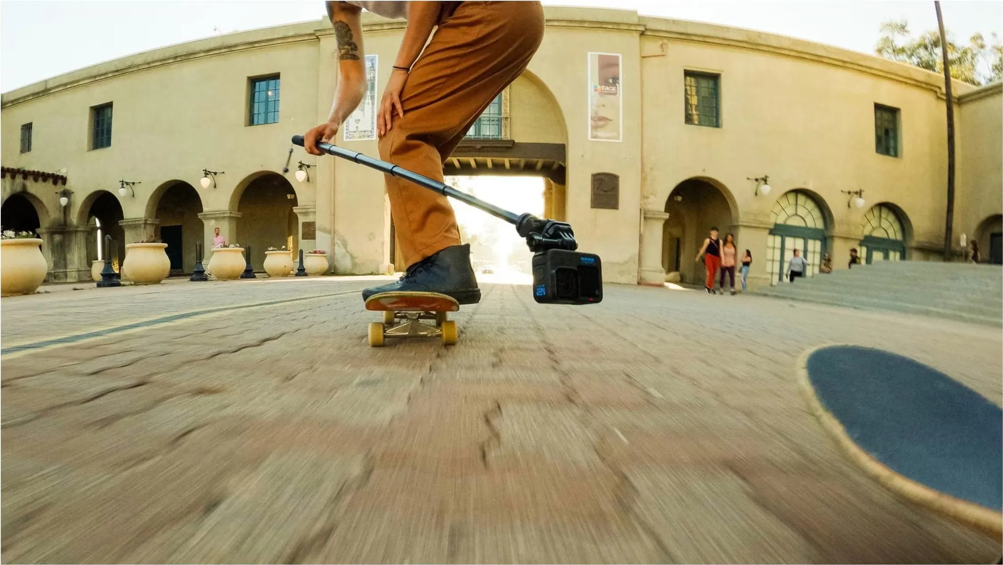 Osoba jeździ na deskorolce, trzyma aparat na kijku selfie. Budynek w tle.