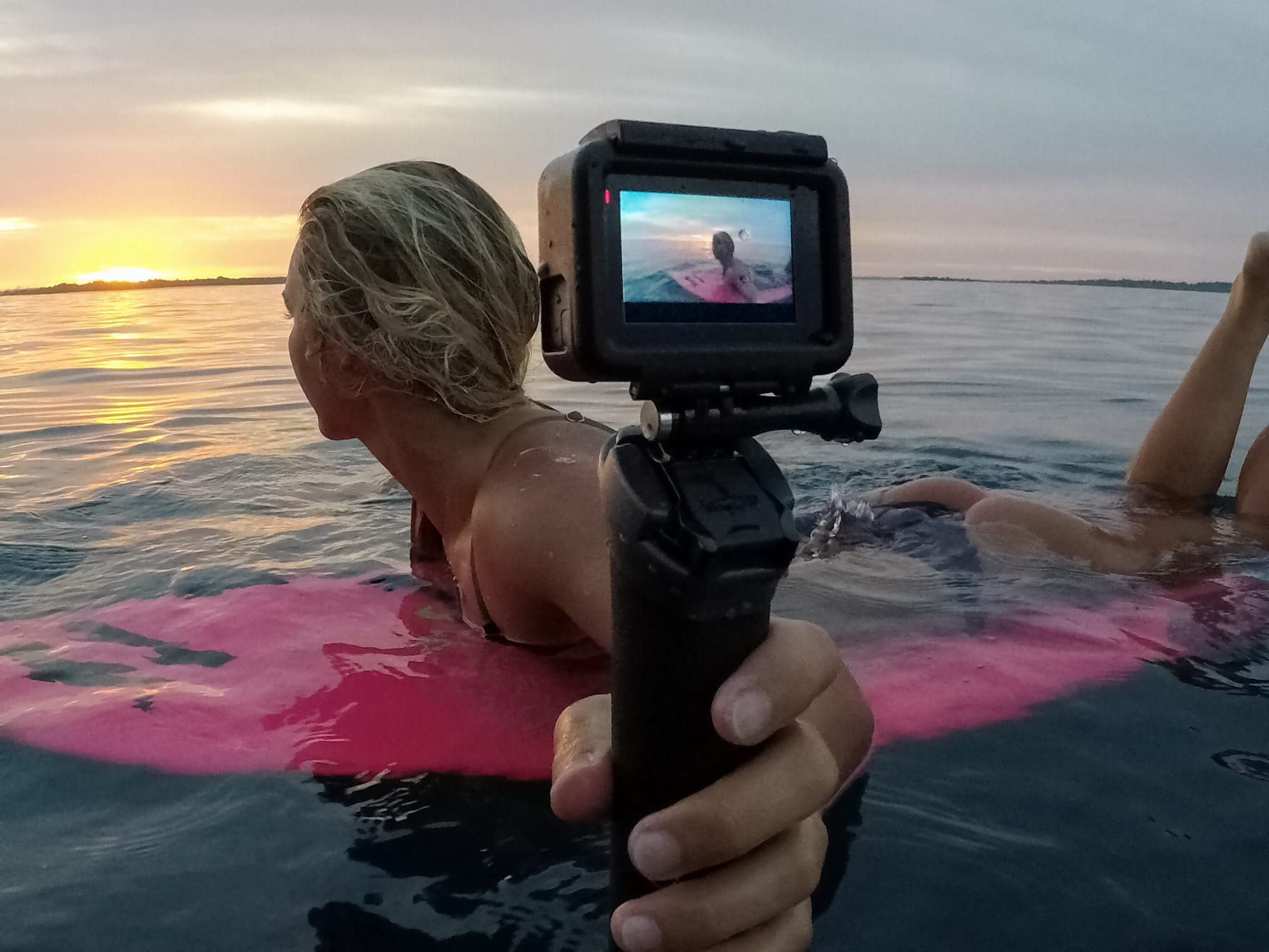 Kobieta surfuje o zachodzie słońca, filmując siebie kamerą GoPro w oceanie.