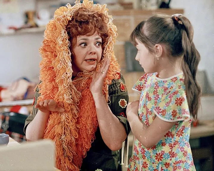 Eine Frau mit orangefarbenem Schal und ein Mädchen im Blumenkleid unterhalten sich.