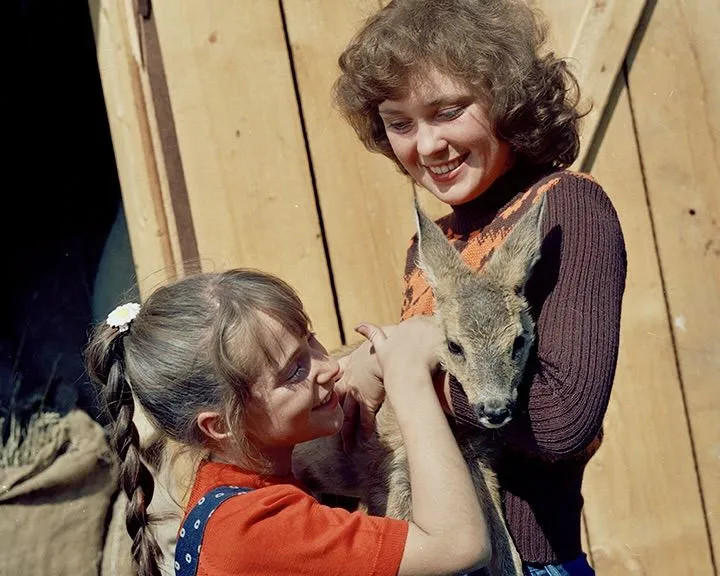 Zwei Mädchen halten ein junges Reh vor einer Holzwand.