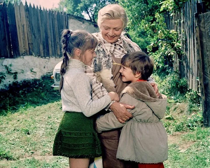 Eine Großmutter umarmt zwei Kinder im Freien vor einem Holzzaun.