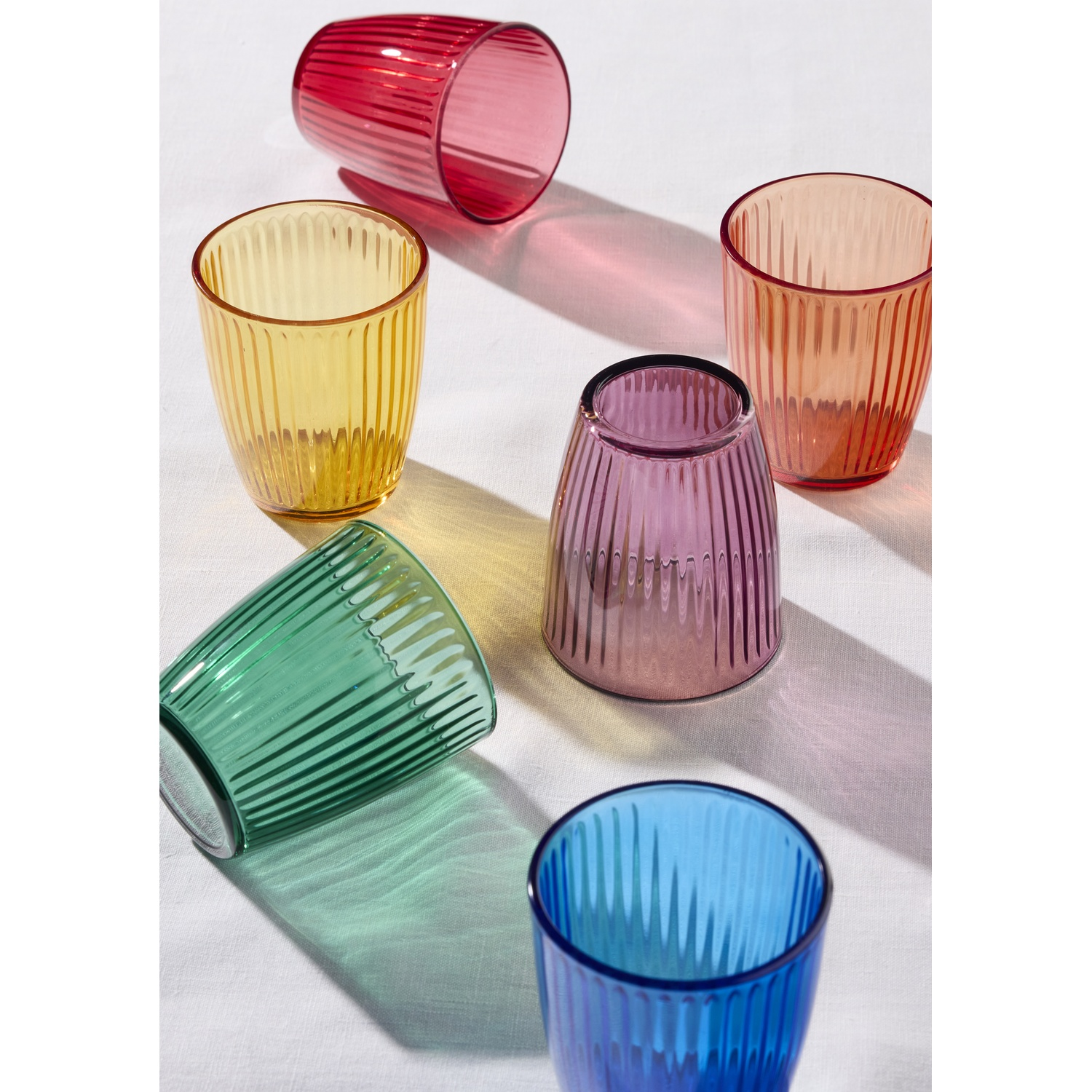 Sei bicchieri colorati a costine disposti su un panno bianco.