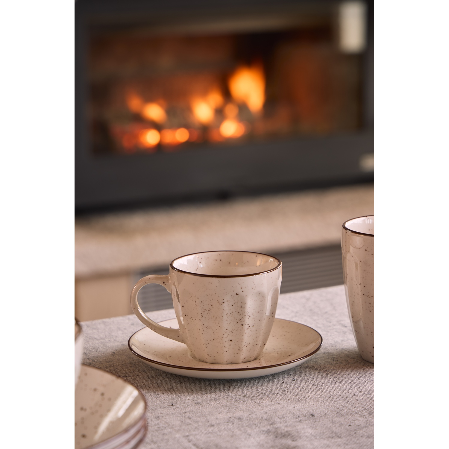Tazza e piattino beige maculati su un tavolo con camino sullo sfondo.