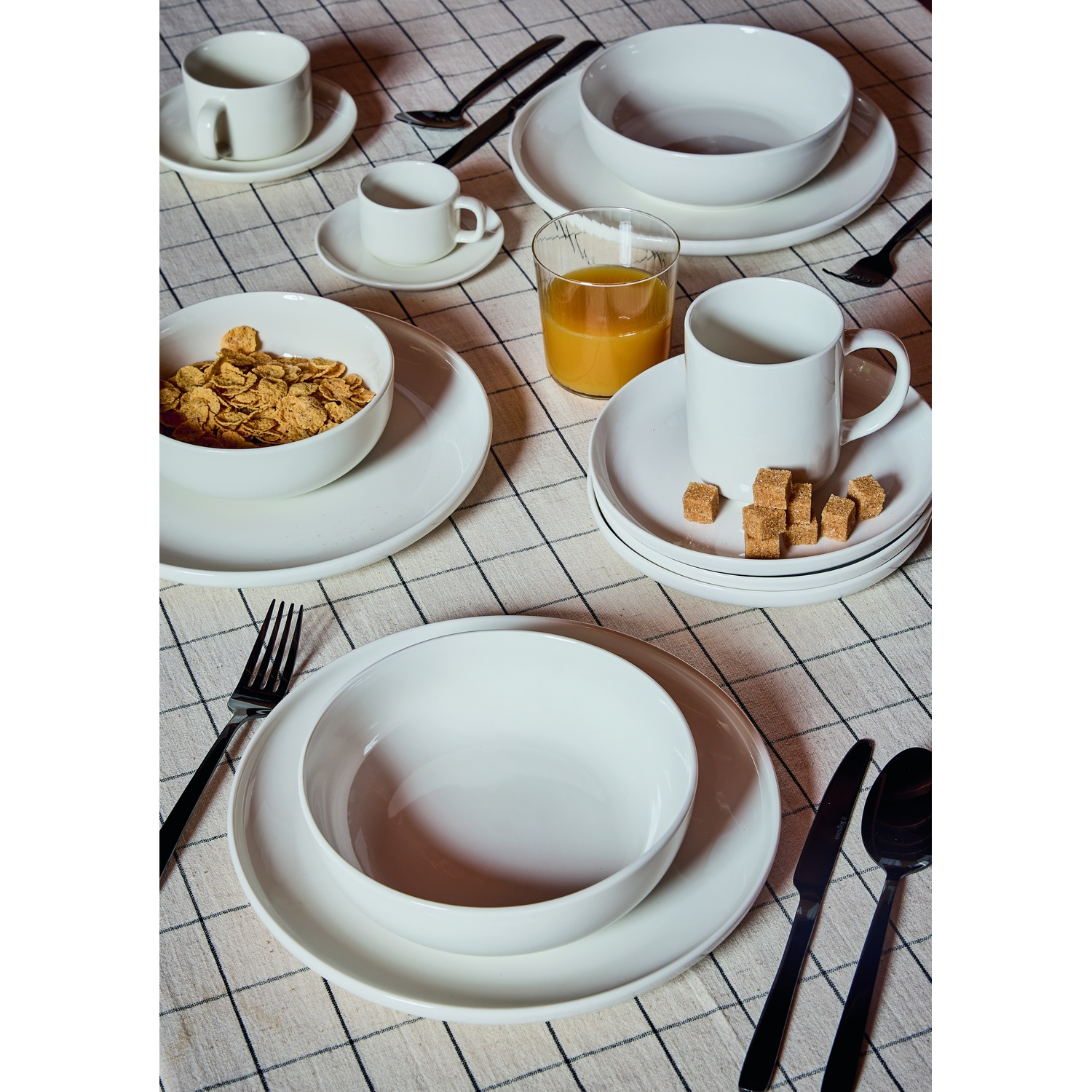 Stoviglie bianche per colazione su tovaglia a quadri: ciotola con cereali, tazze, bicchiere di succo, zollette di zucchero.