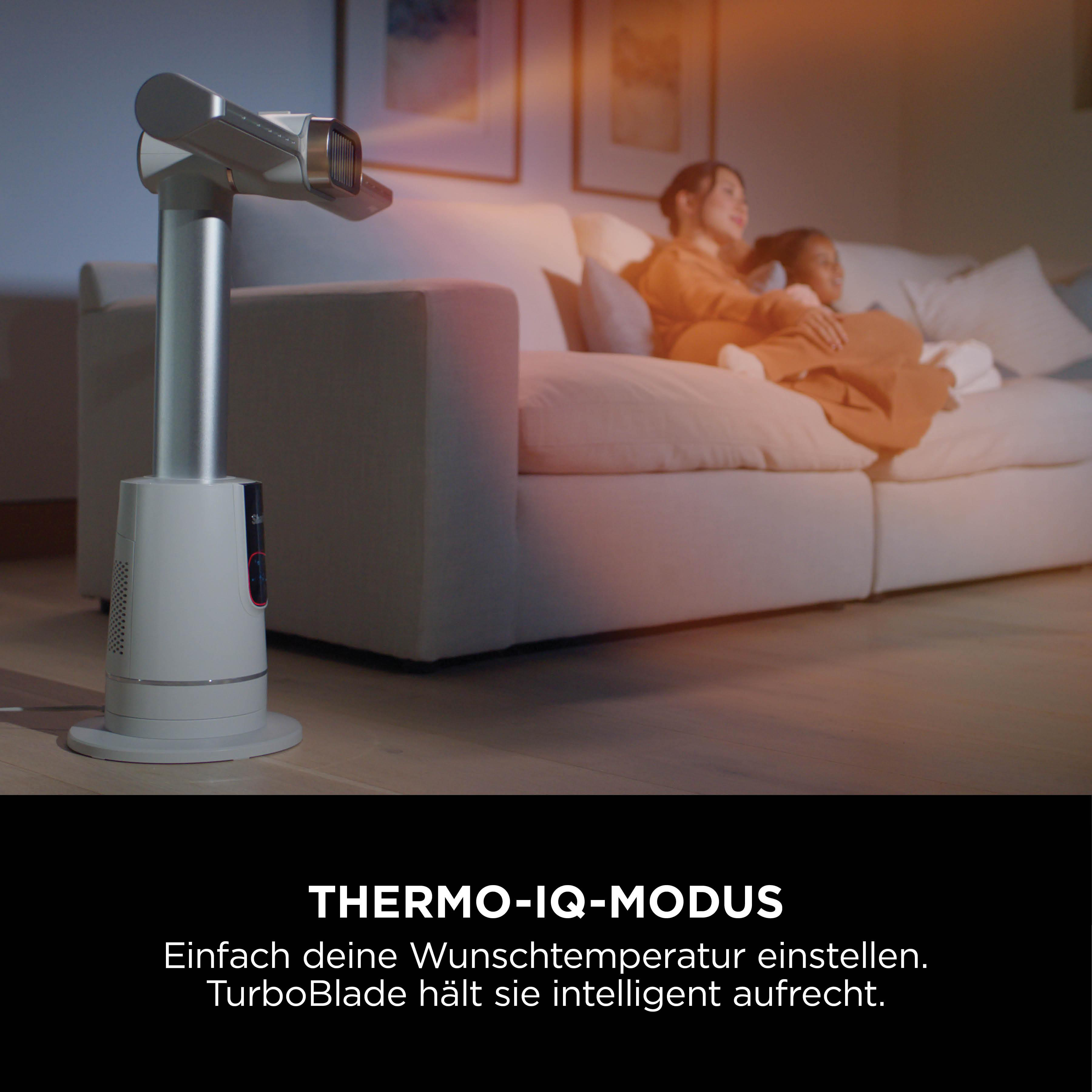 Ein moderner Ventilator steht im Vordergrund, zwei Personen liegen auf einem Sofa im Hintergrund.