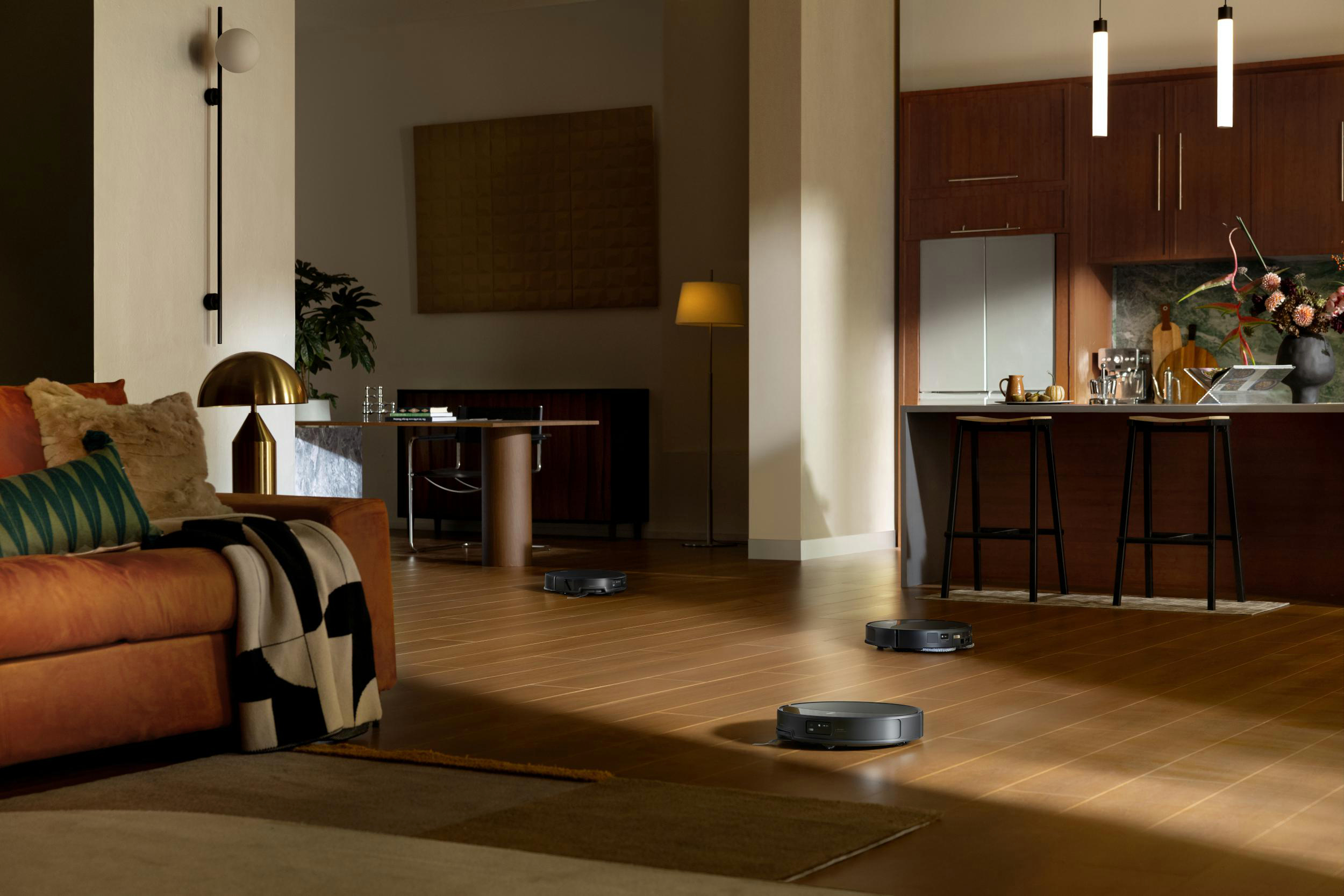 Drei Saugroboter reinigen einen Holzboden in einem modernen Wohnzimmer und einer Küche.