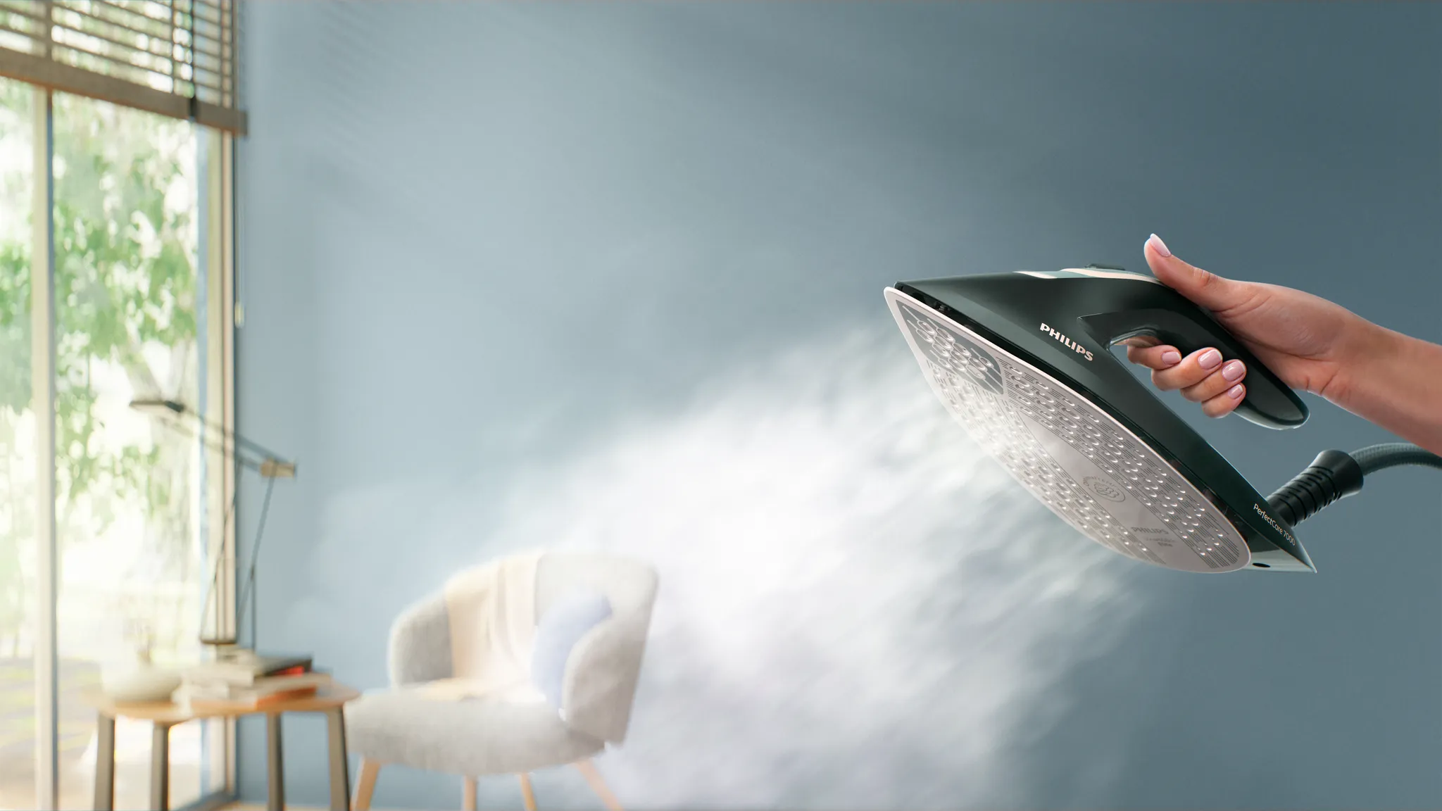 Una mano tiene un ferro Philips nero, che rilascia vapore. Una sedia e un tavolo sullo sfondo.