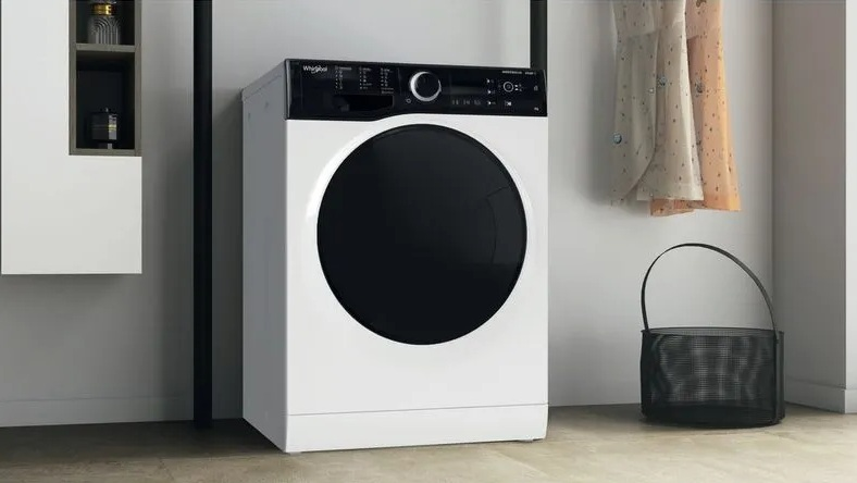 Biała pralka z czarnymi drzwiami i panelem sterowania, obok ubrań i kosza.