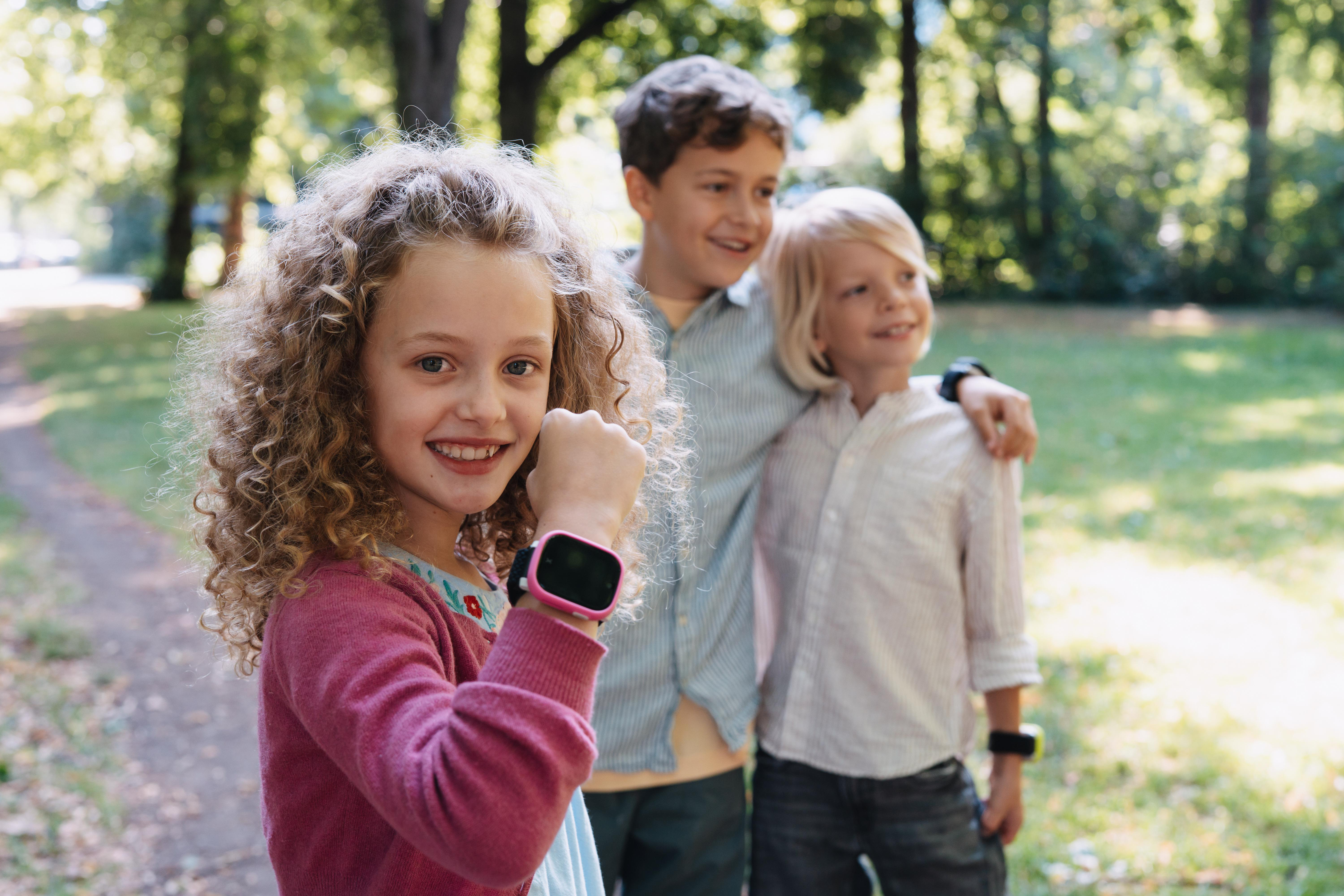 Drei Kinder im Park, ein Mädchen im Vordergrund mit Locken und Smartwatch.