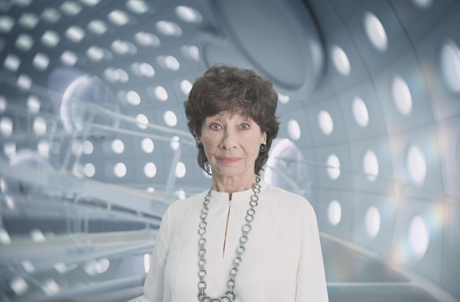 Eine ältere Frau mit kurzen braunen Haaren in einem weißen Oberteil steht vor einem futuristischen Hintergrund.
