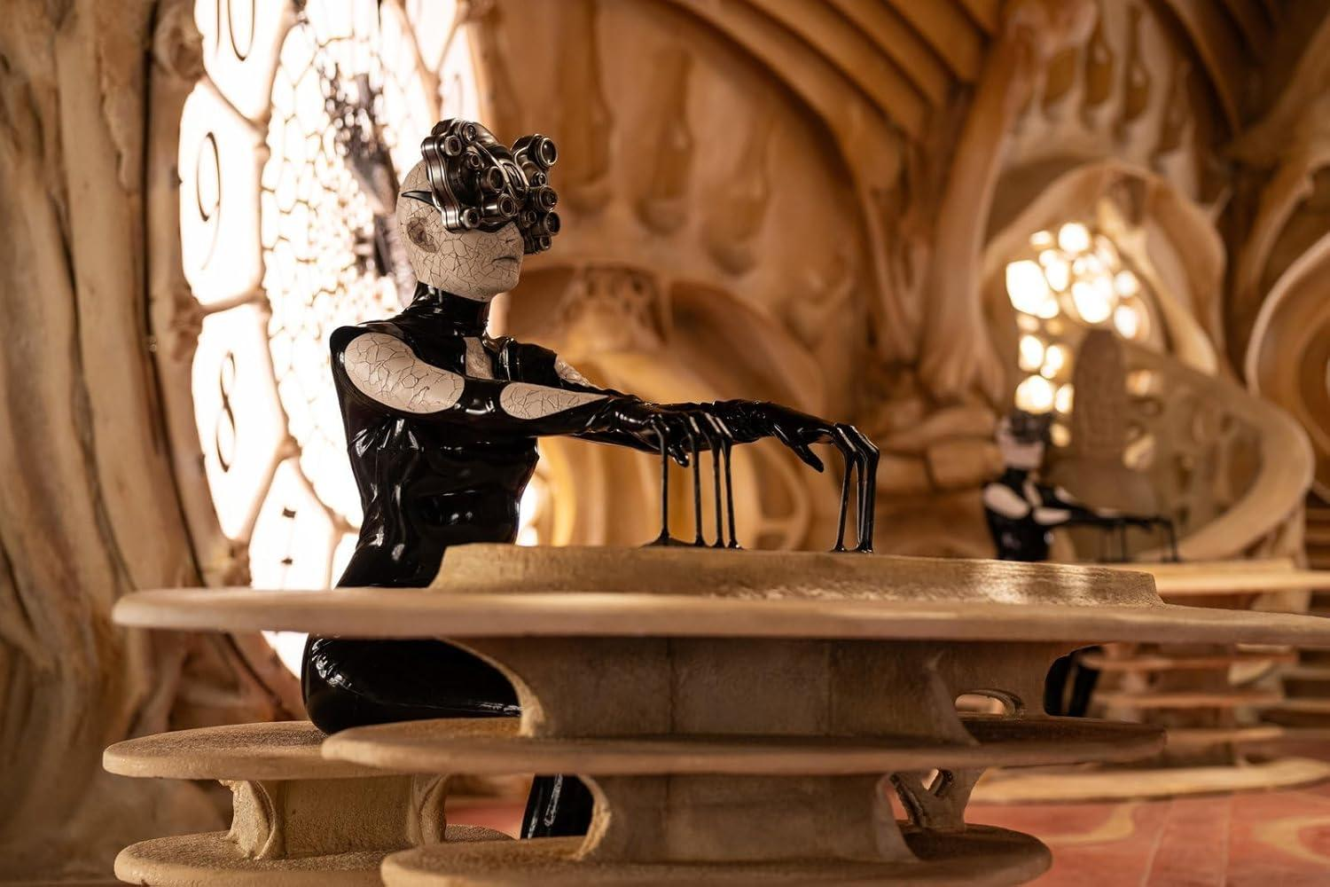Eine Figur in schwarzem Latex sitzt an einem Tisch mit bizarrer Kopfbedeckung.