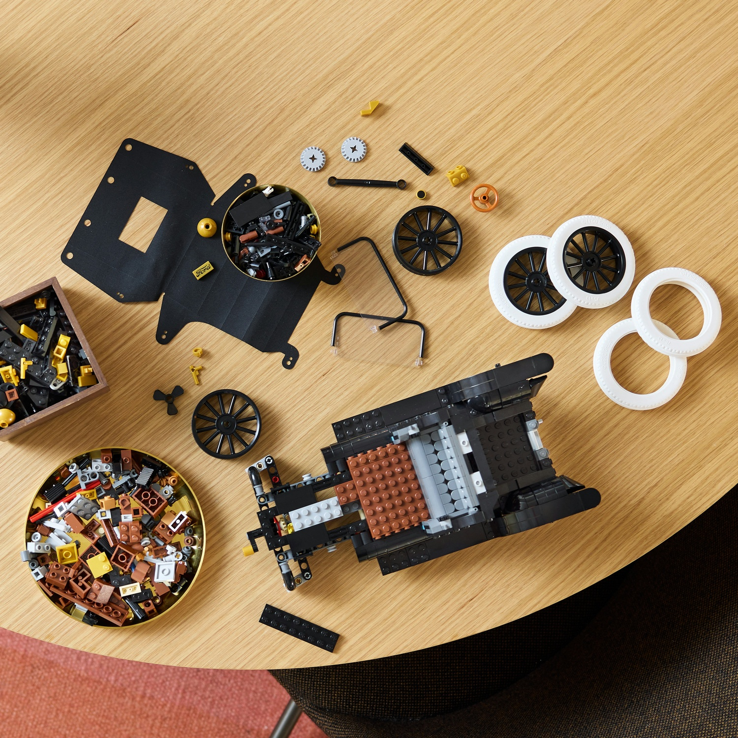 Parti di auto LEGO sparse su un tavolo di legno: telaio nero, ruote e contenitori di mattoncini.