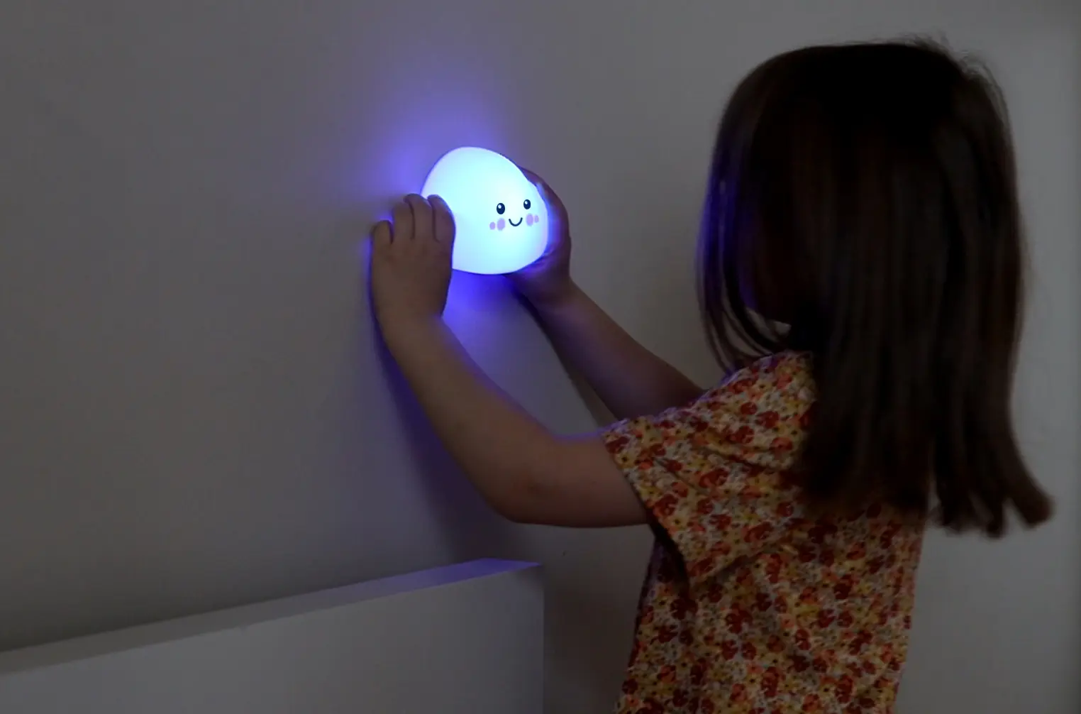 Mała dziewczynka trzyma niebieski, świecący obiekt z twarzą przy ścianie.