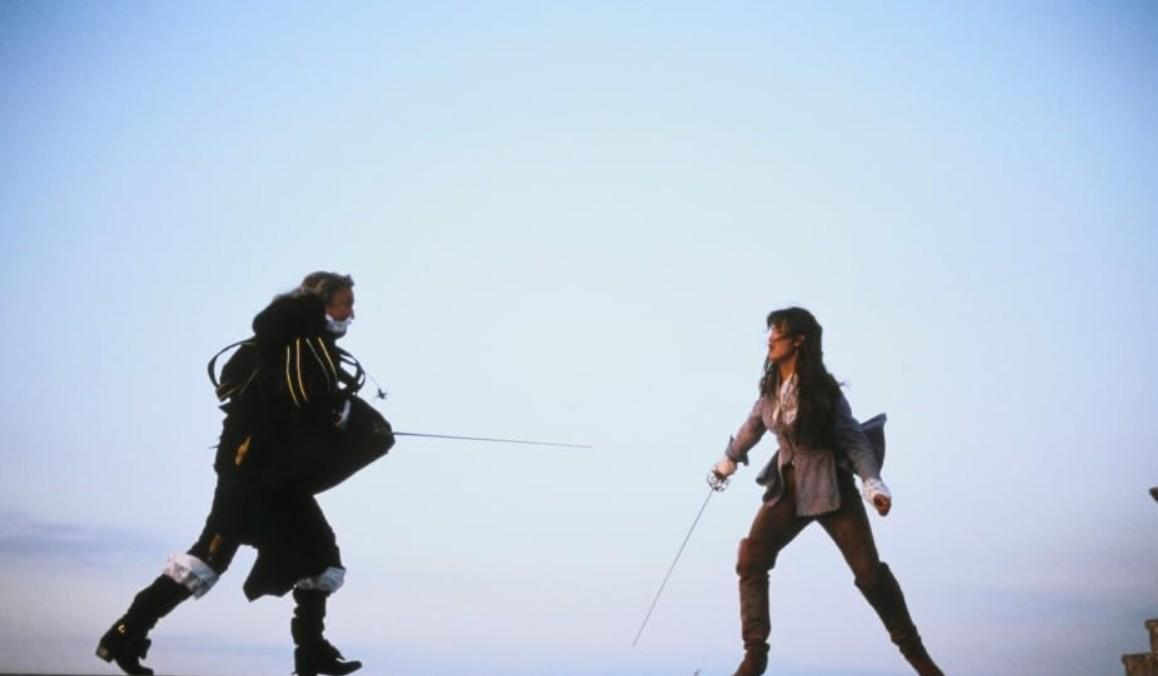 Zwei Fechter duellieren sich vor klarem Himmel. Einer trägt dunkle Kleidung, der andere helle.
