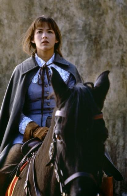 Eine Frau in historischer Kleidung reitet ein Pferd vor einer Steinwand.