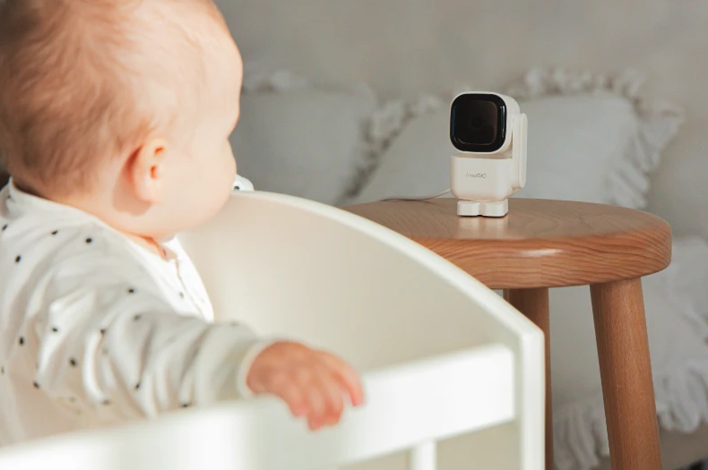 Dziecko siedzi w łóżeczku, patrząc na białą nianię elektroniczną na drewnianym stole.