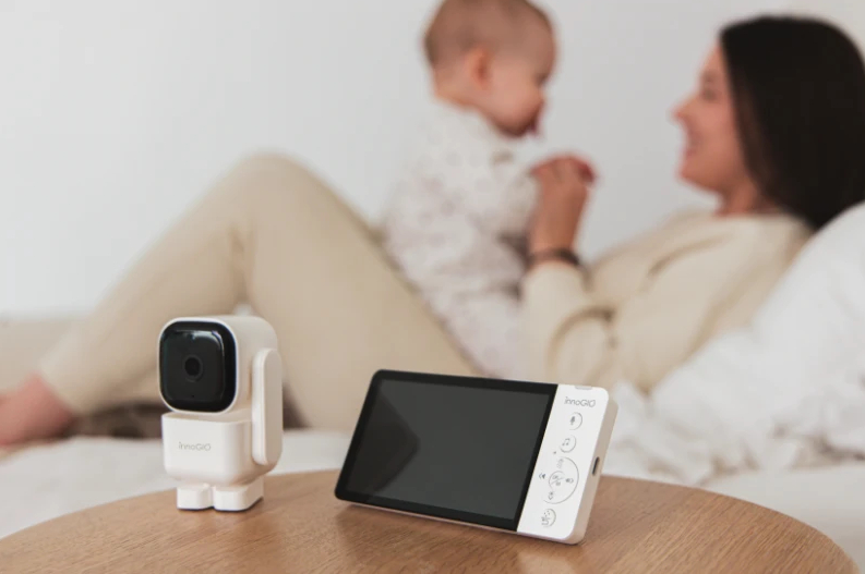 Niania elektroniczna z kamerą i ekranem na drewnianym stole. Kobieta trzyma dziecko w tle.