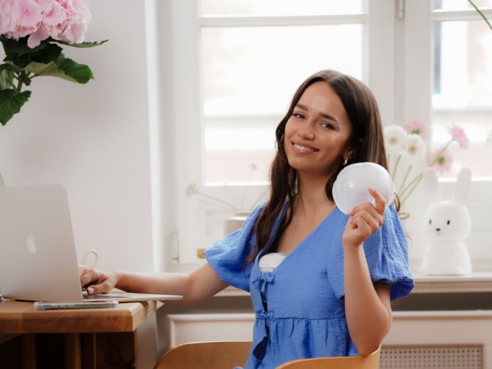 Kobieta w niebieskiej sukience siedzi przy drewnianym biurku z laptopem, trzymając przystawkę do laktatora.
