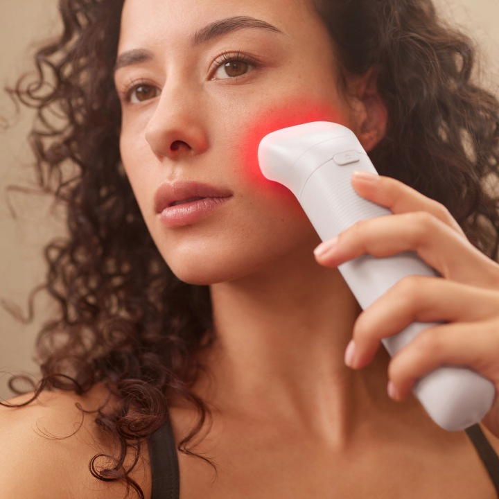 Kobieta z kręconymi włosami używa białego urządzenia do pielęgnacji skóry z czerwonym światłem na policzku.