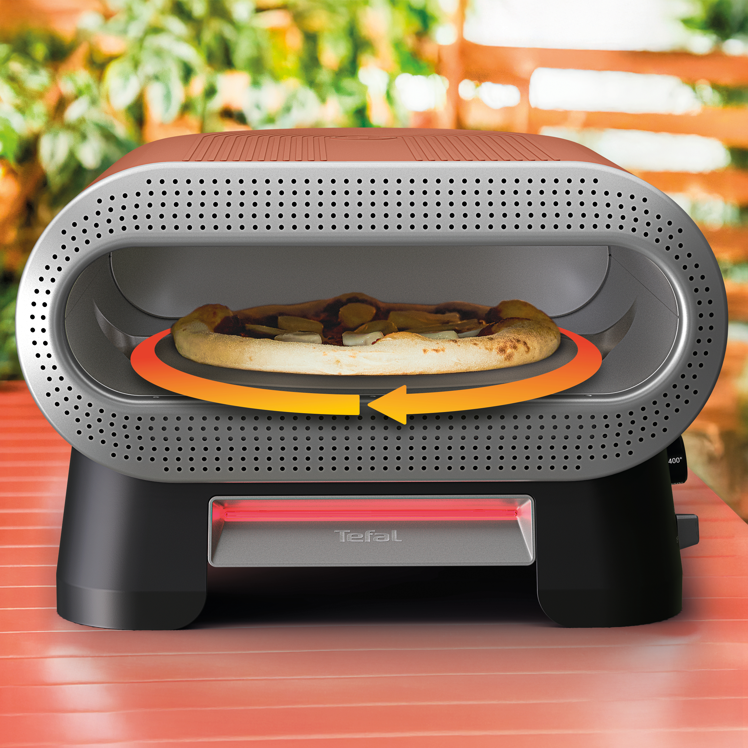 Eine Pizza dreht sich in einem Tefal-Ofen auf einem roten Tisch, mit grüner Vegetation im Hintergrund.