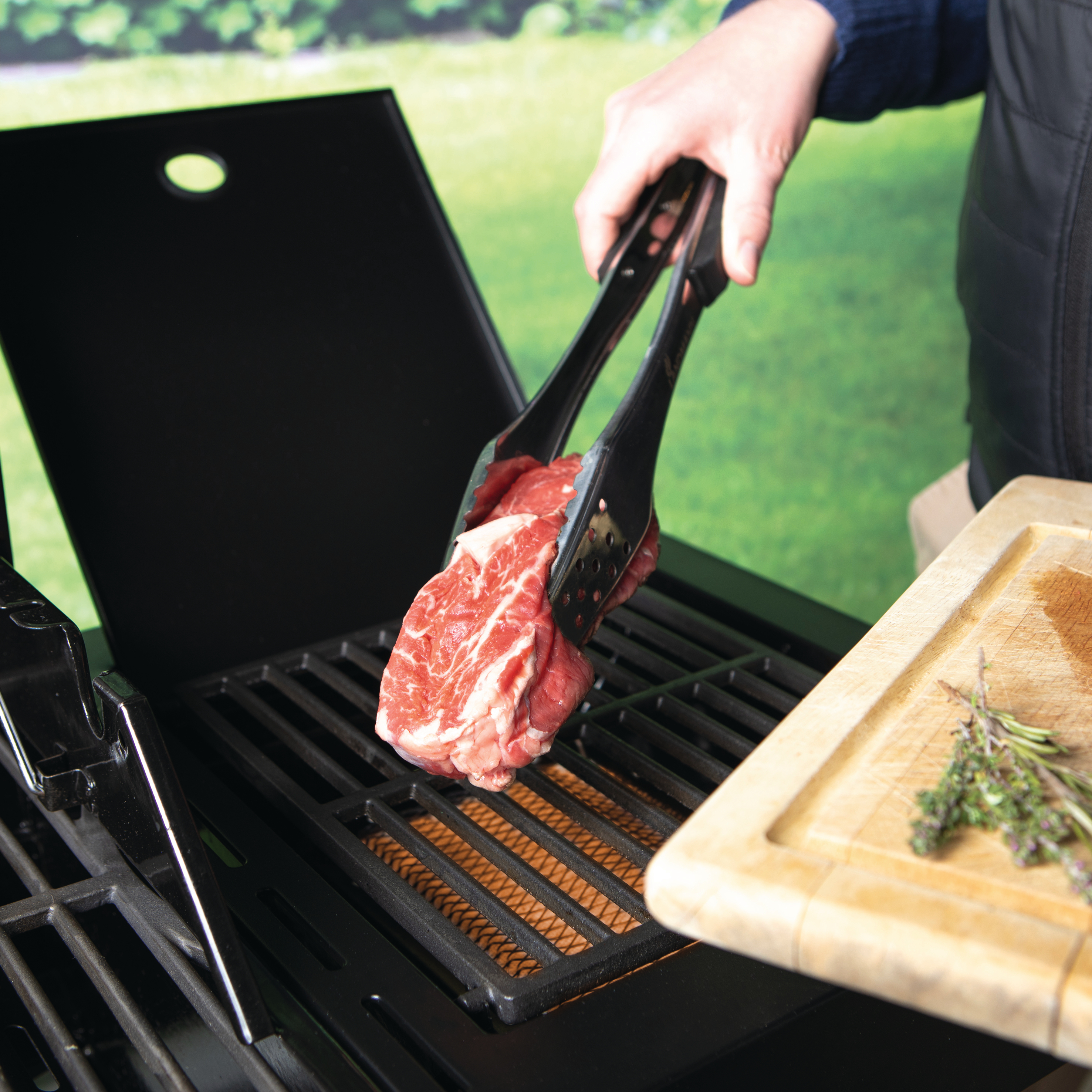 Fleisch wird mit Zange auf einen Grill gelegt, draußen im Grünen.