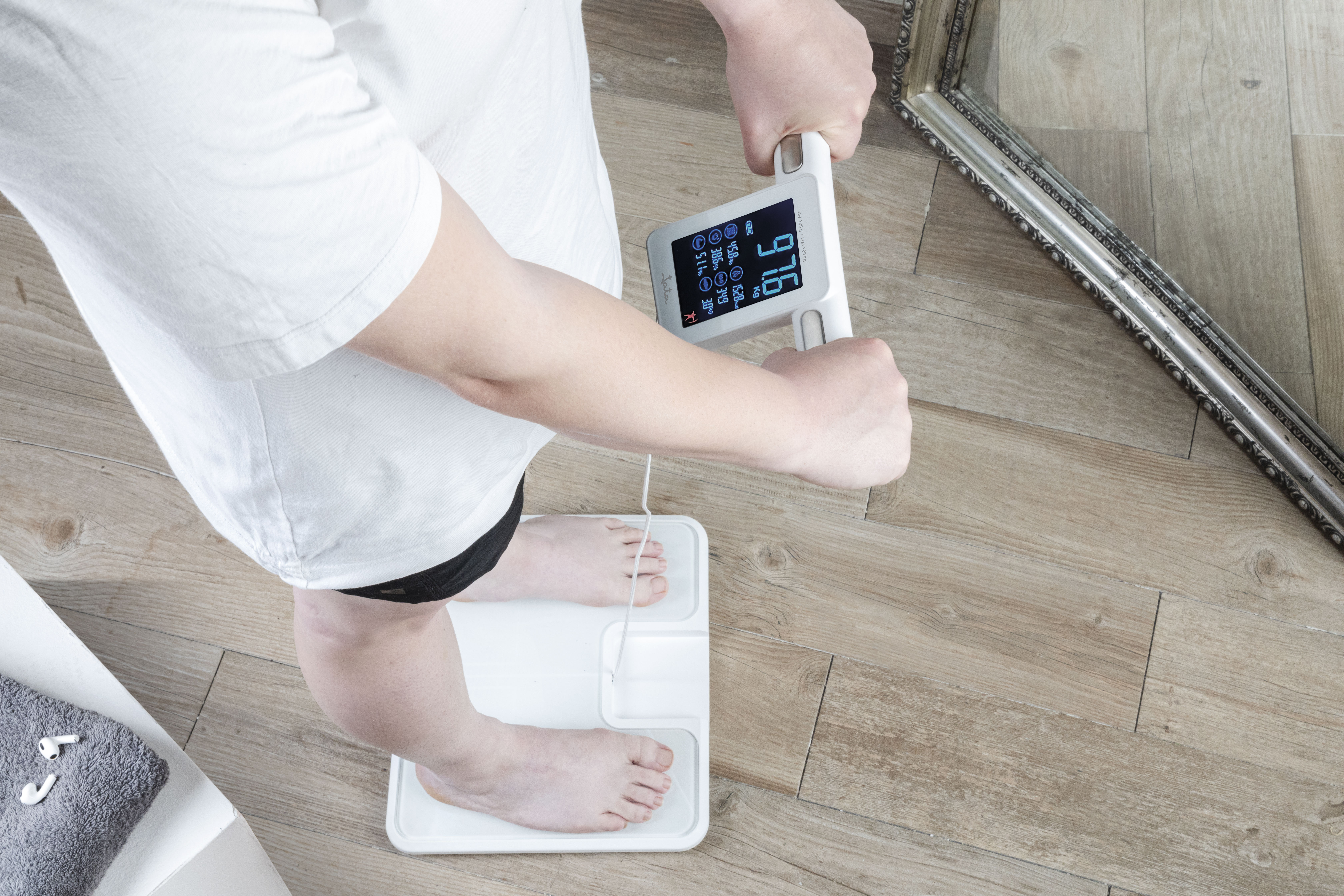 Una persona con camisa blanca está en una báscula blanca, sosteniendo un dispositivo inteligente. La báscula marca 97.6 kg.