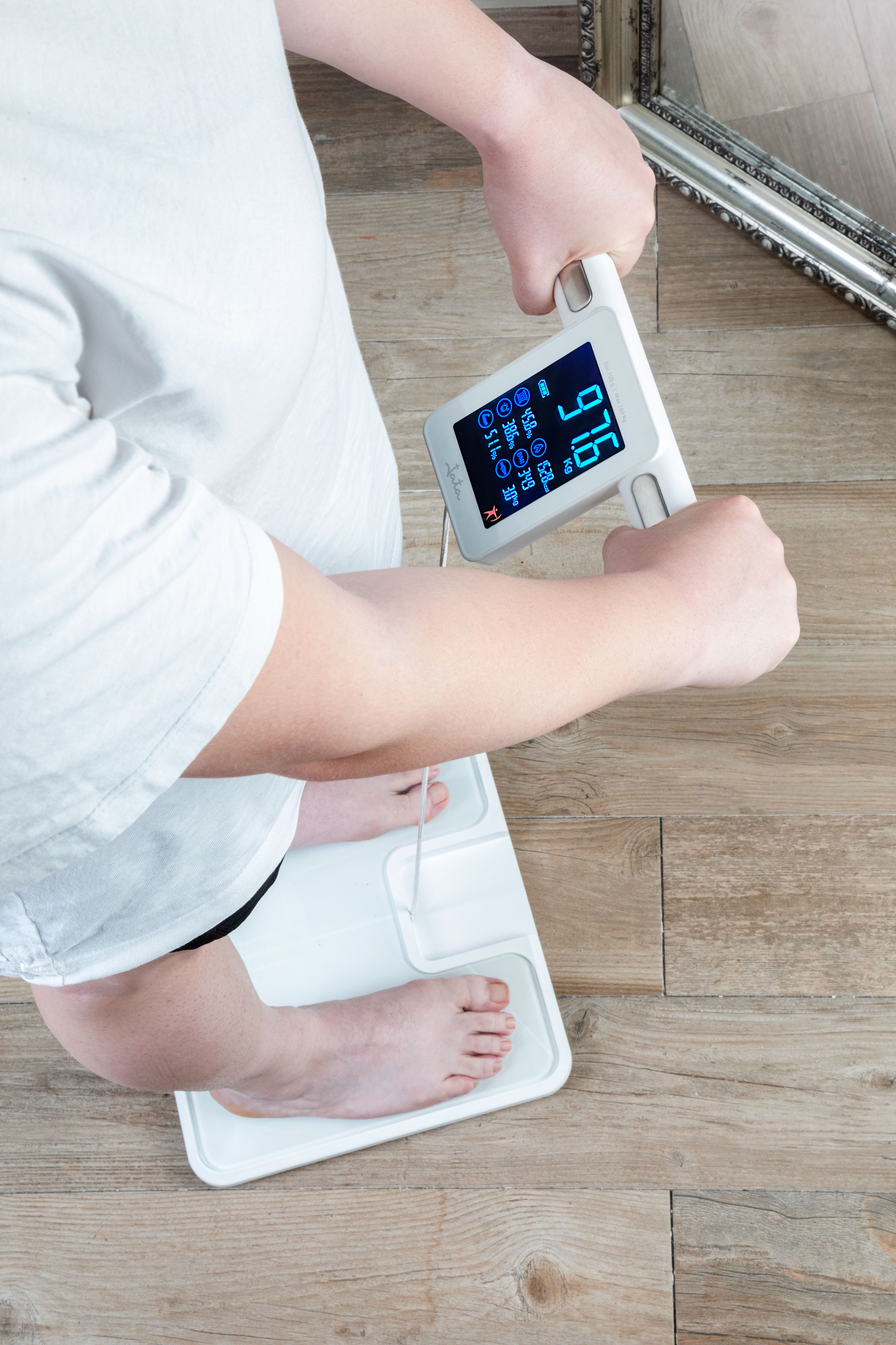 Una persona se para en una báscula blanca. La pantalla muestra 97,6 kg.