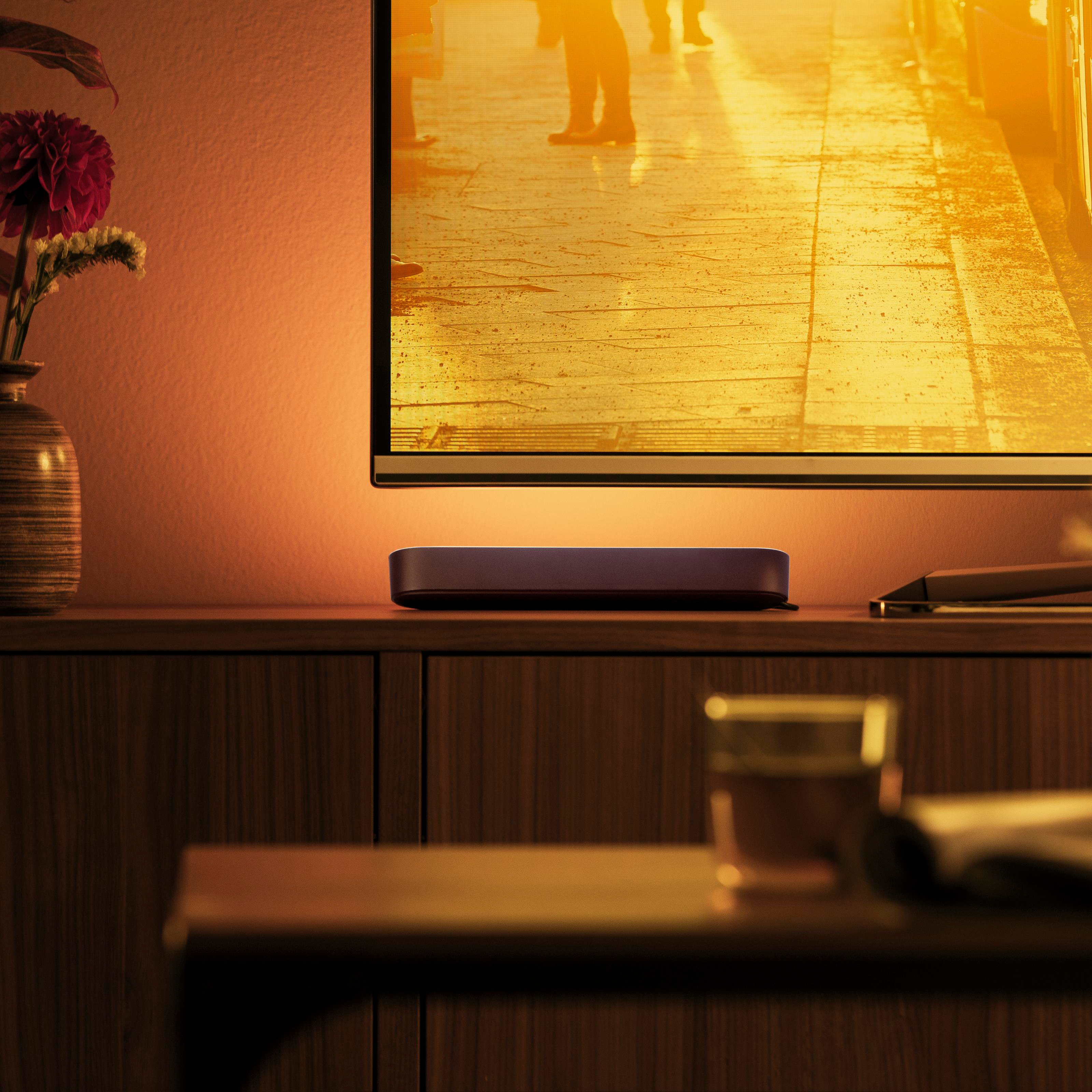 Ein roter Blume in einer Vase neben einem Fernseher mit orangefarbener Beleuchtung.