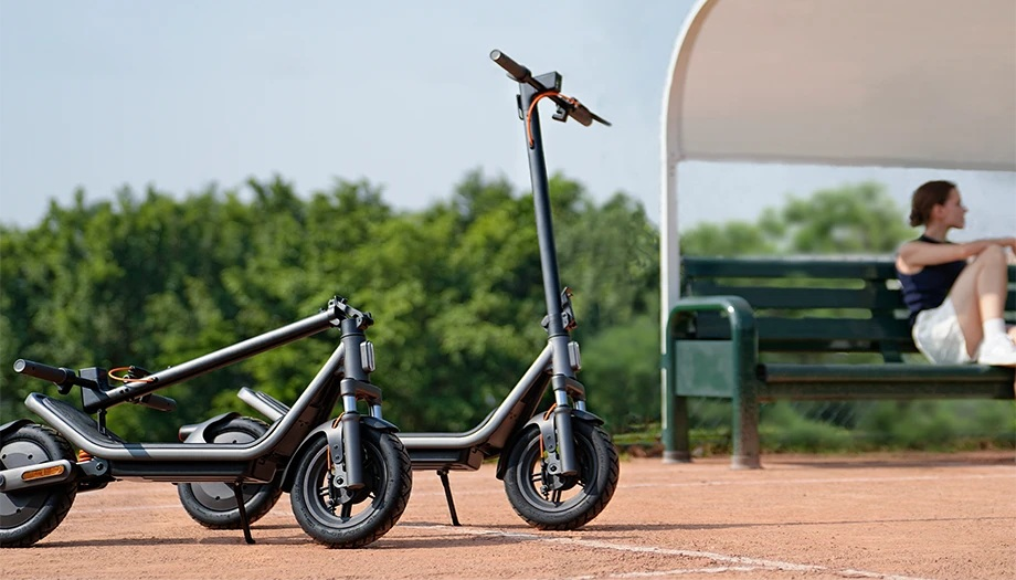 Dwa ciemne hulajnogi elektryczne stoją na czerwonym korcie. Kobieta siedzi na ławce w tle.